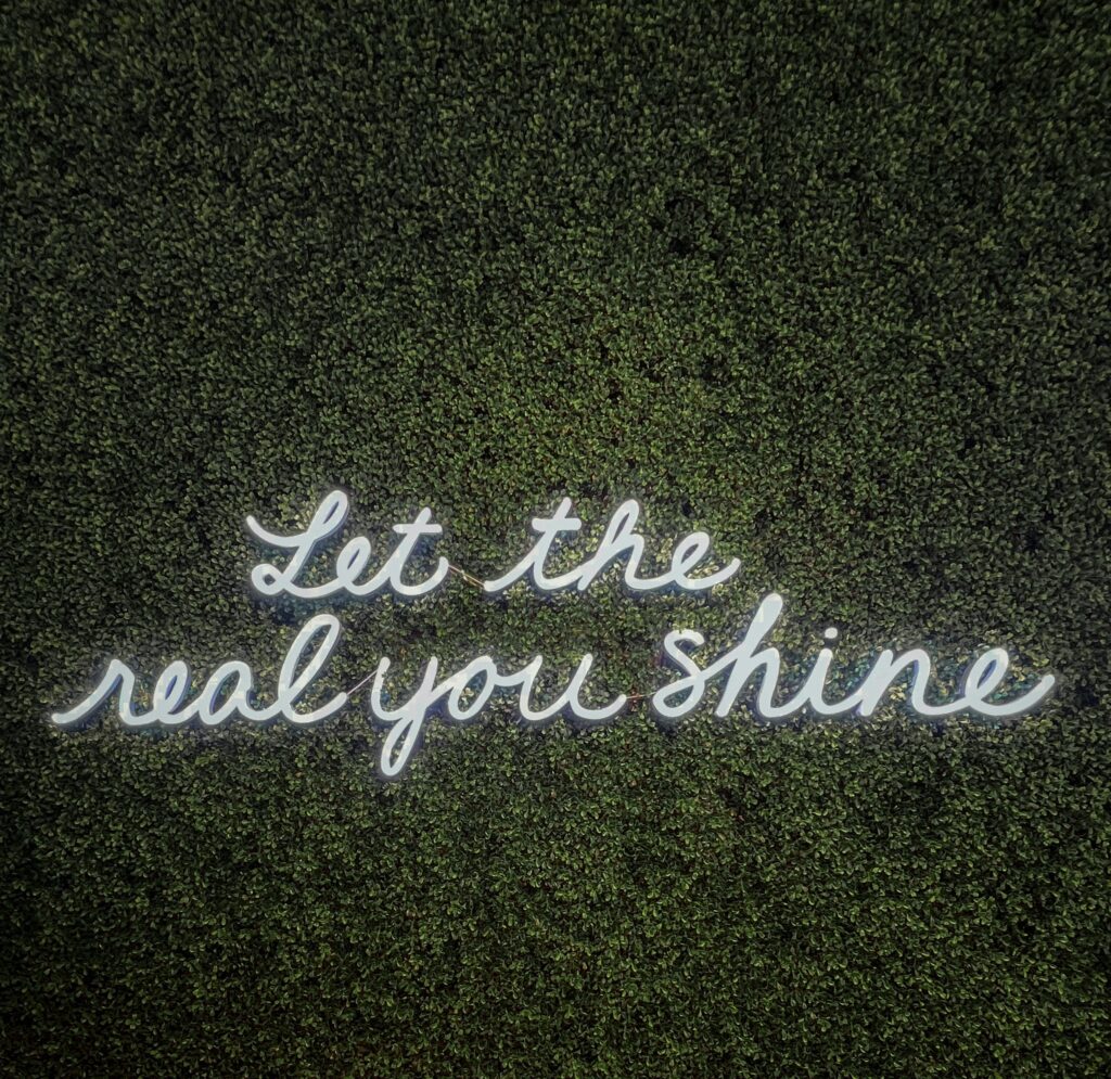 Phrase en anglais, "let the real you shine", écrite avec des néons en forme de lettres sur un mur végétal.